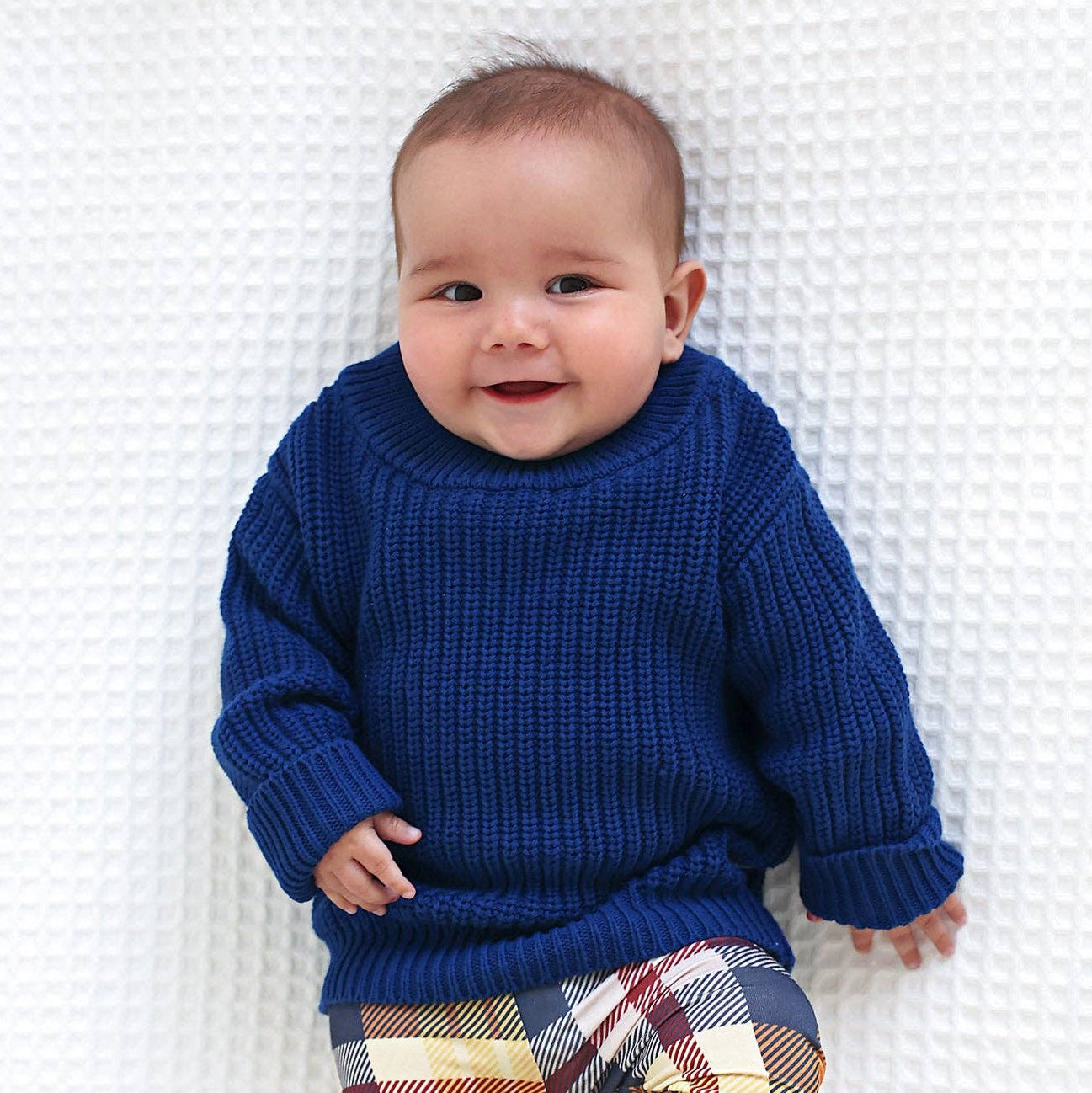 Baby wearing a blue sweater and plaid pants on a white blanket